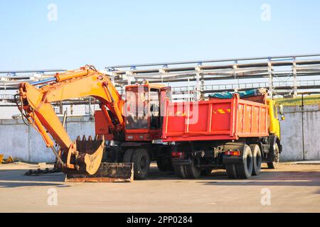 Un grande camion giallo arancione pesante con un rimorchio, un camion di scarico e un escavatore con una siviera sono parcheggiati in fila in un cantiere edile. Foto Stock