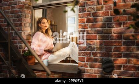 Bella Donna in Pink Sweater è sorridente, digitando sul laptop e seduto sul suo davanzale. Blogger femminile scrivendo un articolo sul benessere Foto Stock
