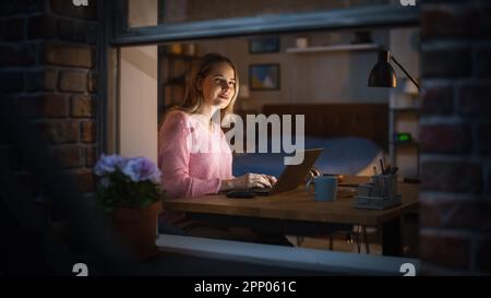 Bella donna bionda lavora su un computer portatile mentre si siede alla sua scrivania in camera da letto. Freelancer professionista femminile che lavora da casa accogliente di notte. Foto Stock