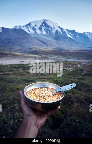 Recipiente turistico pieno di tagliatelle fast food e cucchiaio in mano nella natura montagna valle campeggio nel trekking dell'Asia centrale, Kazakistan Foto Stock