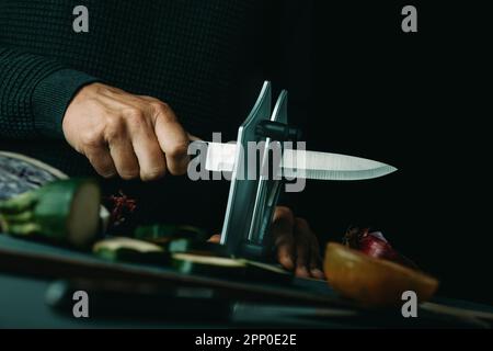 l'uomo affina un coltello da cucina con un affilatore da tavolo su un tavolo con alcune verdure tagliate e tritate Foto Stock