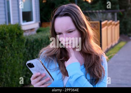 Giovane donna bionda riccia elegante scrive messaggio sullo smartphone a piedi sul parco vicino al fiume, hipster turista in viaggio in città europea e pianificazione di viaggio utilizzando il telefono cellulare. Ragazza indossare maglione Foto Stock