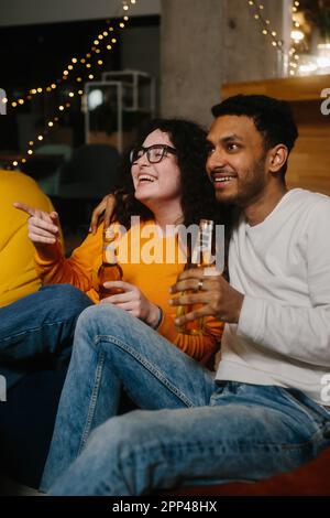 Un paio di amici multirazziali stanno guardando un film comico mentre si siedono su borse di fagioli morbidi mentre bevono birra. Foto Stock