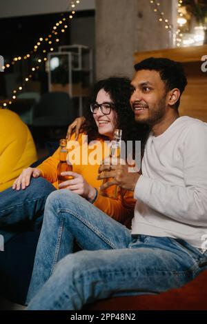 Un paio di amici multirazziali stanno guardando un film comico mentre si siedono su borse di fagioli morbidi mentre bevono birra. Foto Stock