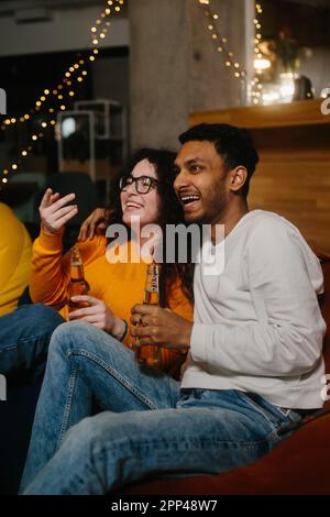 Un paio di amici multirazziali stanno guardando un film comico mentre si siedono su borse di fagioli morbidi mentre bevono birra. Foto Stock