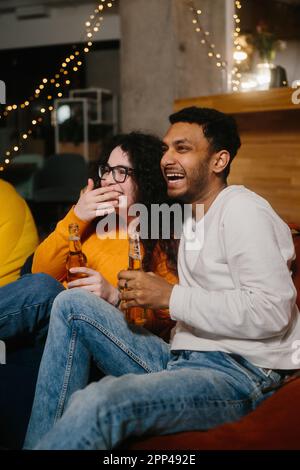 Un paio di amici multirazziali stanno guardando un film comico mentre si siedono su borse di fagioli morbidi mentre bevono birra. Foto Stock