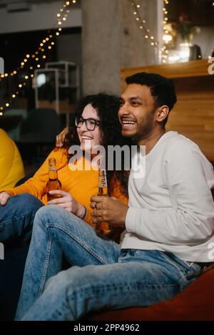 Un paio di amici multirazziali stanno guardando un film comico mentre si siedono su borse di fagioli morbidi mentre bevono birra. Foto Stock