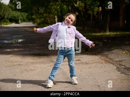 Ritratto a tutta lunghezza di adorabile bambina con raccolta giallo dente di leone fiore, sorridendo guardando la macchina fotografica, esprimendo emozioni positive, godere Foto Stock