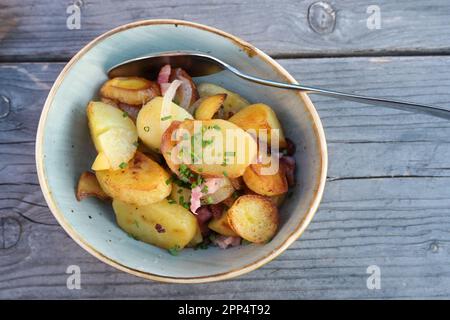 Patate fritte con cipolle, pancetta e contorno di erba cipollina in una ciotola di ceramica su un tavolo di legno grigio blu, vista ad angolo alto dall'alto, spazio copia, selezionato fo Foto Stock