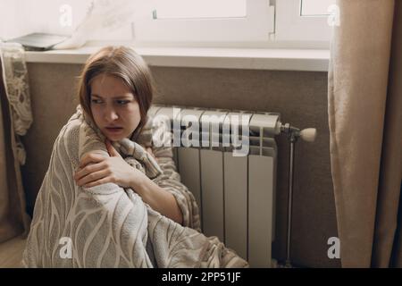 Una donna sofferente in un plaid si siede alla batteria di riscaldamento e si riscalda in casa e si congela. Foto Stock