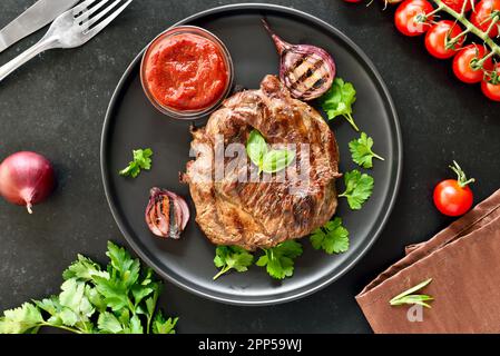 Succosa bistecca di manzo alla griglia e salsa di pomodoro sul piatto su fondo di pietra nera. Vista dall'alto, disposizione piatta Foto Stock