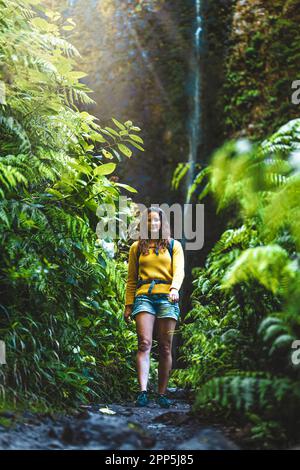 Descrizione: Backpacker donna a piedi lungo felce percorso cresciuto a pittoresca, cascata in crescita nella foresta pluviale di Madeiran. Levada di Caldeirão V Foto Stock