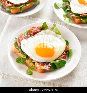 Apri i sandwich con uova fritte, pancetta e foglie di verdure sul piatto su sfondo di pietra chiaro. Vista ravvicinata Foto Stock