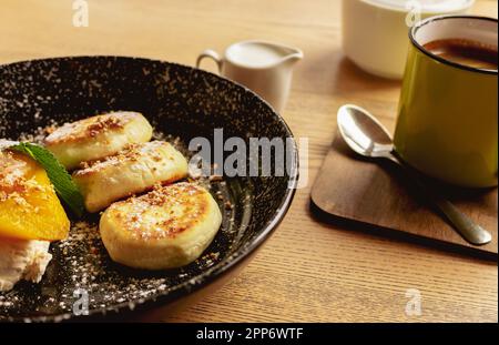 Syrniki con pera al caramello e tazza da caffè. Frittelle cagliate con caffè e latte. Colazione deliziosa. Cibo ucraino. Frittelle al formaggio e caffè. Foto Stock