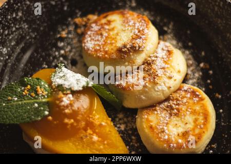 Syrniki con pera al caramello e ricotta. Frittelle cagliate su piatto nero. Colazione deliziosa. Cibo ucraino. Pera al forno con foglia di menta e frittella Foto Stock