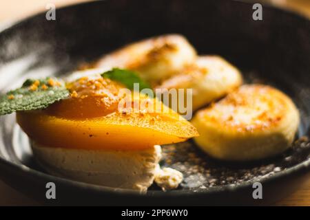 Syrniki con pera al caramello e ricotta. Frittelle cagliate su piatto nero. Colazione deliziosa. Cibo ucraino. Pera al forno con foglia di menta e frittella Foto Stock