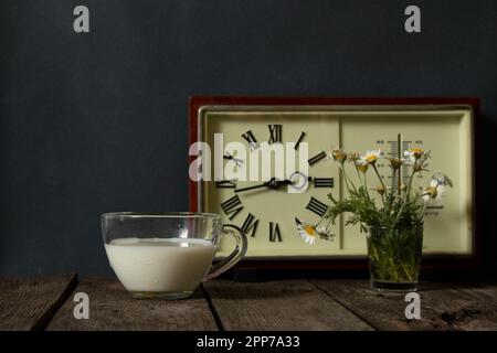 una tazza di latte e un vaso con margherite vicino all'orologio su un tavolo di legno Foto Stock