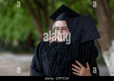 I laureati caucasici dell'European College e neri che indossano cappellini e abiti abbracciano dopo la laurea, orgogliosi del successo accademico, dei diplomi e della conoscenza Foto Stock