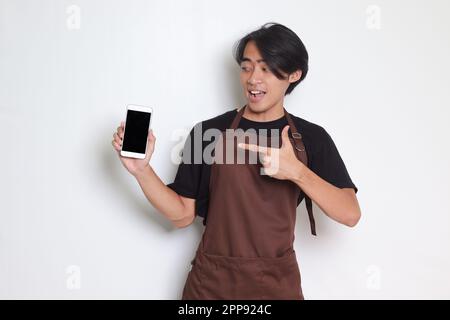 Ritratto dell'attraente barista asiatico in grembiule marrone che mostra lo schermo vuoto del telefono cellulare. Spazio vuoto per la simulazione. Isolato su sfondo bianco Foto Stock