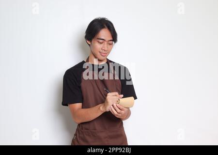 Ritratto dell'attraente barista asiatico in grembiule marrone che prende ordine, scrivendo sulla lista del libro del menu. Immagine isolata su sfondo bianco Foto Stock