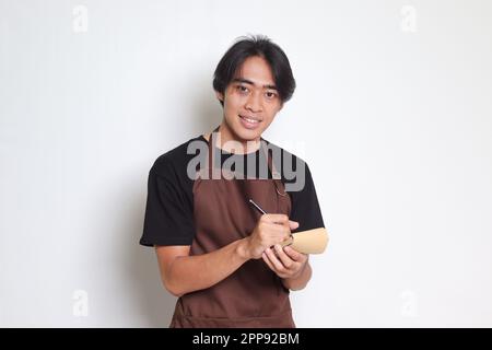 Ritratto dell'attraente barista asiatico in grembiule marrone che prende ordine, scrivendo sulla lista del libro del menu. Immagine isolata su sfondo bianco Foto Stock