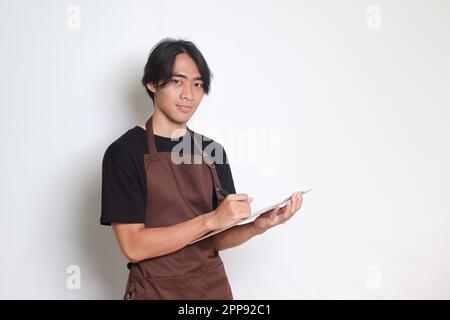 Ritratto dell'attraente barista asiatico in grembiule marrone che prende ordine, scrivendo sulla lista del libro del menu. Immagine isolata su sfondo bianco Foto Stock