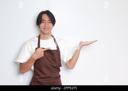 Ritratto dell'attraente barista asiatico in grembiule marrone che mostra il prodotto, indicando qualcosa con le mani. Concetto pubblicitario. Immagine isolata su cui Foto Stock