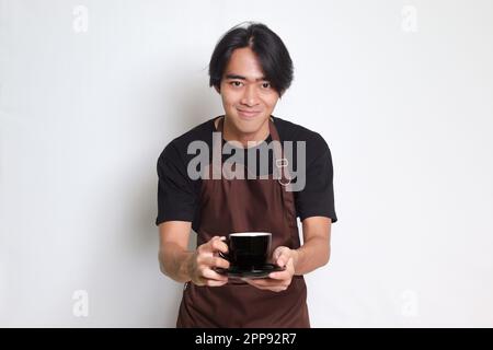 Ritratto dell'attraente barista asiatico in grembiule marrone che serve una tazza di caffè al cliente. Immagine isolata su sfondo bianco Foto Stock