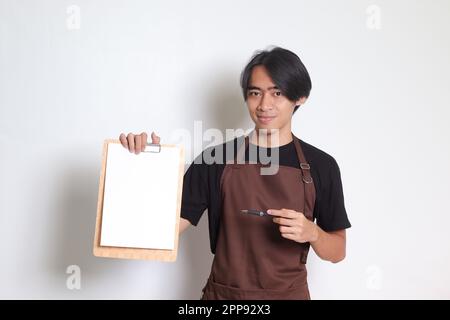 Ritratto dell'attraente barista asiatico in grembiule marrone con carta bianca del menu sulla clipboard. Immagine isolata su sfondo bianco Foto Stock
