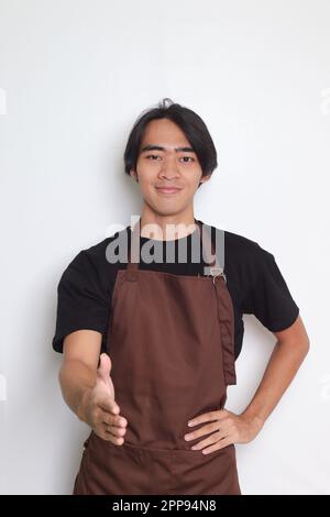 Ritratto dell'attraente barista asiatico in grembiule marrone che si protesta per una stretta di mano e saluta qualcuno. Immagine isolata su sfondo bianco Foto Stock