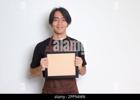 Ritratto dell'attraente barista asiatico in grembiule marrone con bordo a cornice vuoto. Immagine isolata su sfondo bianco. Foto Stock