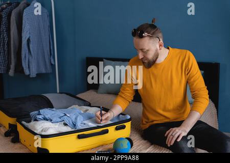 Uomo millenario che fa un check-list di cose da portare a casa. Valigia da imballaggio a casa. Concetto di viaggio Foto Stock