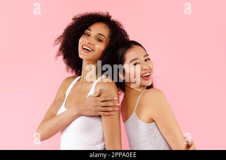 Due attraenti donne afro-americane e asiatiche che posano su sfondo rosa studio, guardando e sorridendo alla macchina fotografica Foto Stock