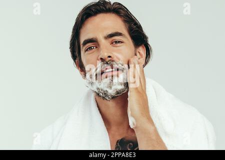 Giovane uomo che applicava la crema da barba alla barba, preparandosi a una rasatura confortevole e liscia. Uomo che pratica la sua routine di governare, prendendosi cura Foto Stock