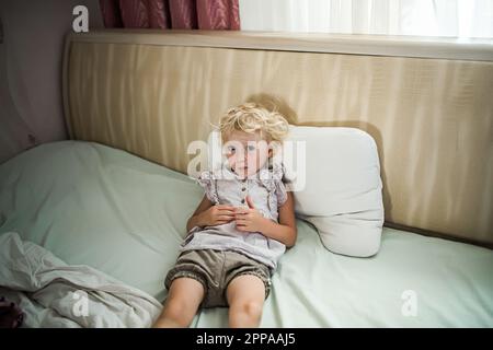 piccola ragazza triste si trova su letto largo incrociate le mani e si rifiuta di dormire. Emozioni ed esperienze dei bambini. Foto Stock