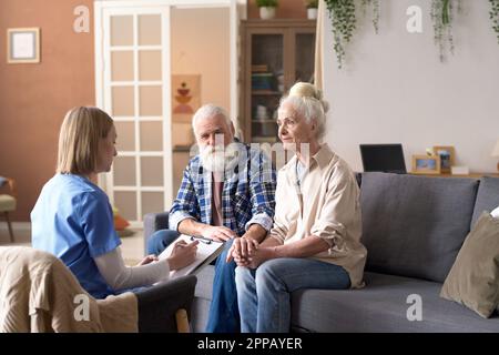 Giovane infermiera che chiede informazioni sulle condizioni di salute a coppie anziane e facendo appunti in tessera medica durante la sua visita a casa Foto Stock