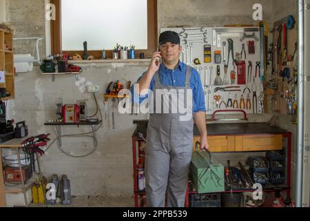 Immagine di un handyman nel suo workshop che parla al telefono e tiene in mano una cassetta degli attrezzi. Risposta a una chiamata per lavoro. Foto Stock