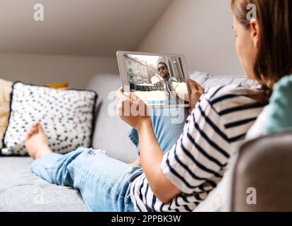 Uomo che viaggia e chiama la sua ragazza in video. Donna sdraiata sul divano a casa e in chat con il suo ragazzo online utilizzando un tablet digitale. Foto Stock