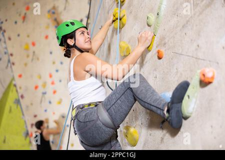 Ben attrezzato, positivo allenamento giovane donna su muro di pietra, montagna artificiale con rocce, arrampicata in parco bouldering al coperto Foto Stock