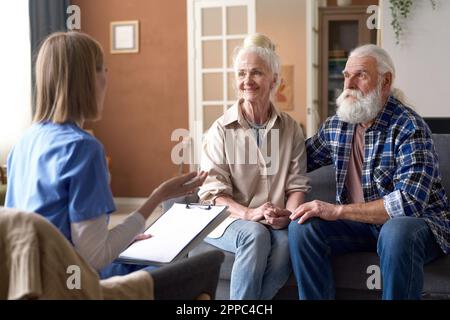 Giovane infermiera che parla con la coppia anziana e che prende appunti in tessera medica durante la sua visita a casa Foto Stock