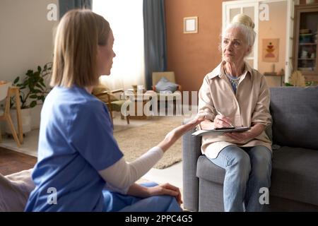 Giovane infermiera che parla con una donna anziana mentre la ascolta e prende appunti seduti sul divano a casa Foto Stock