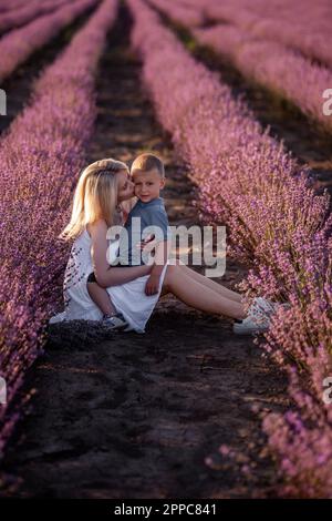 Ritratto di madre bionda con figlio piccolo sono seduti in campo di lavanda viola. Giovane donna in abito bianco abbraccia e bacia amorevolmente il ragazzo. La conce Foto Stock