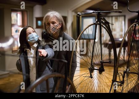 Tween studentessa e anziana donna tutor in maschere di viso guardando la bicicletta d'epoca nel museo Foto Stock