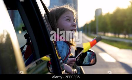 Primo piano di bella bambina tenendo in mano la bandiera tedesca e guardando fuori dalla finestra dell'auto con il volto stupito. Bambino piccolo carino che viaggia in Germania sopra Foto Stock