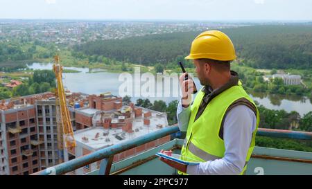 Ritratto di un bel costruttore uomo caucasico in uniforme e casco guardando il tablet digitale e parlando in walkie-talkie mentre si è in piedi Foto Stock