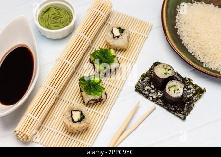 Sushi di alghe impostato su un tappeto di sushi di bambù. Wasabi e salsa di soia in ciotole. Riso su un piatto. Sushi con cetriolo su foglio di nori. Giacitura piatta. Sfondo bianco Foto Stock