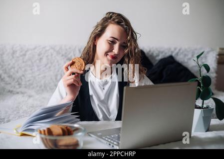 Giovane donna redhead guardando serie o podcast guardando lo schermo con interesse tenere cookie in mano seduto al tavolo da cucina con vaso di vetro e. Foto Stock