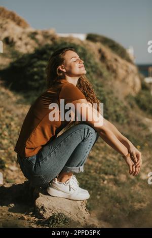 Donna con gli occhi chiusi accovacciandosi sulla roccia durante il giorno di sole Foto Stock