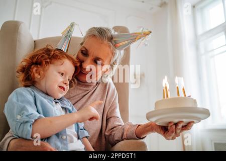 Donna anziana con nipote che tiene la torta di compleanno a casa Foto Stock
