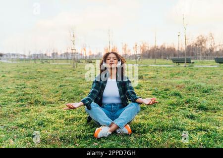 Giovane donna che indossa cuffie wireless meditando nel parco Foto Stock